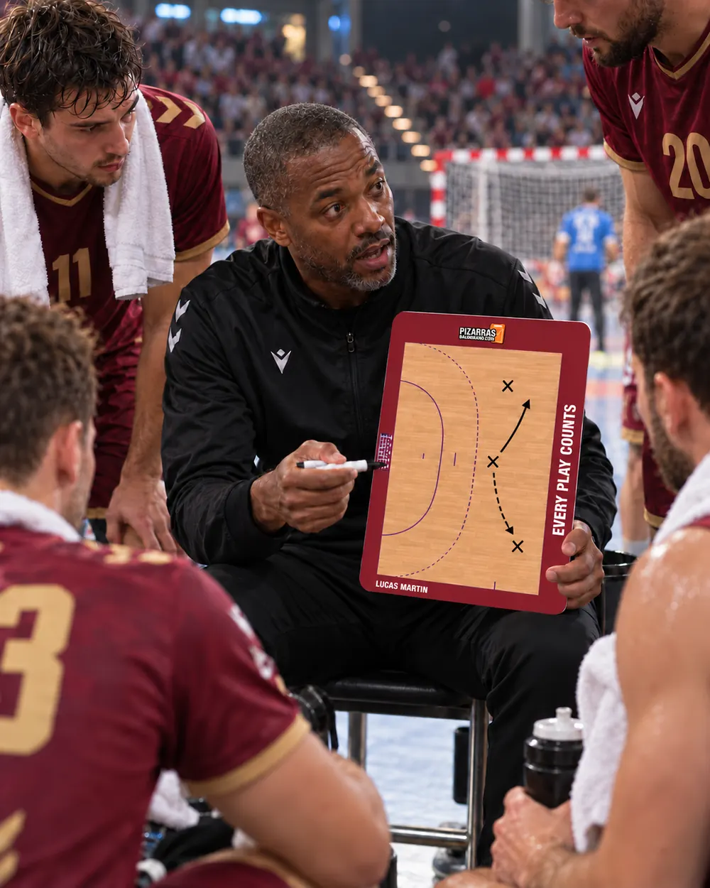 A handball coach explains a play on the whiteboard to his players in a packed arena