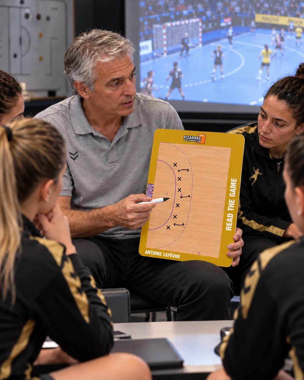 A handball coach explains a play on the whiteboard to his players in front of the video screen